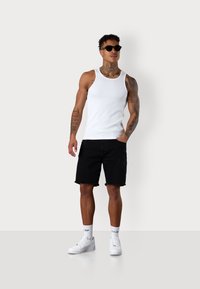 White ribbed tank top, black denim shorts with frayed hem, white sneakers, and sunglasses. Model has tattoos and a straight posture.