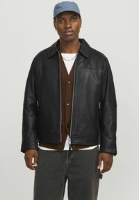 Black leather jacket with a collared neckline, zippered front, side pockets, and a relaxed fit, worn over a brown cardigan and white shirt.