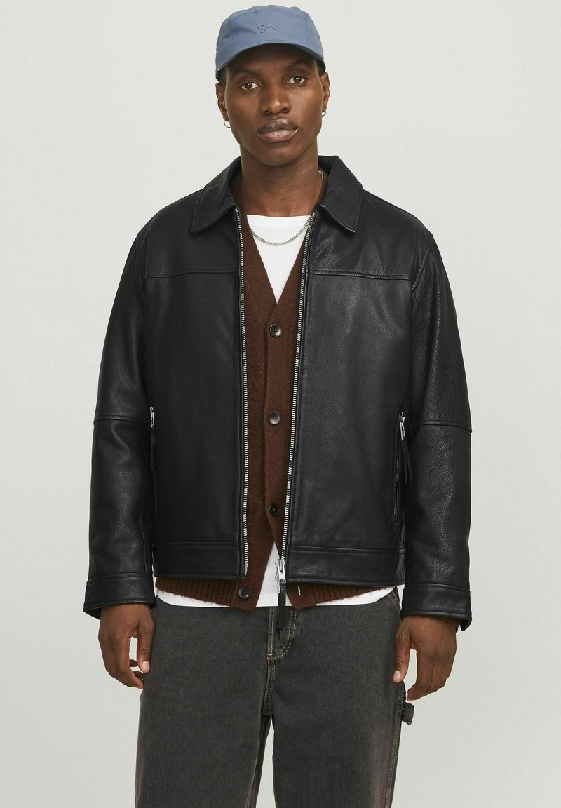 Schwarze Lederjacke mit einem Kragen, Reißverschluss vorne, seitlichen Taschen und lässiger Passform, getragen über einem braunen Cardigan und einem weißen Hemd.