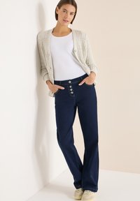 Vaqueros azul marino con cintura ajustada y bragueta de cinco botones, combinados con un cárdigan gris claro texturizado y una camiseta blanca.