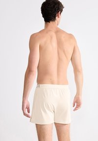 Ljusbeiga herrboxershorts med elastisk midja, med en liten logotyp, sedd bakifrån utan ytterligare kläder.