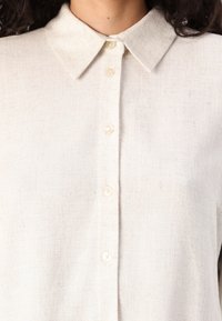 Lys beige lin skjorte med kragehals, fem knapper foran og strukturvev. Har subtile variasjoner i farge og tekstur.