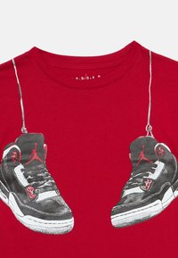 Red shirt featuring a graphic of black, white, and red Air Jordan sneakers hanging by their laces around the neckline.