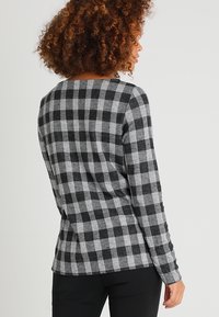 Femme aux cheveux bouclés portant une chemise à manches longues à carreaux noirs et gris, vue de dos sur un fond uni.