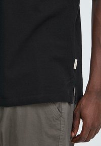 Seitenansicht einer Person, die ein schwarzes Shirt mit einem kleinen "BLEND"-Label und graue Hosen mit sichtbaren Nähten nahe der Tasche trägt, entspannte Hand.