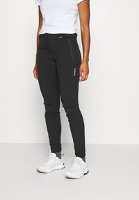 Sorte strækbukser med slim fit, lynlåslommer og en tekstureret overflade, parret med hvide sneakers.