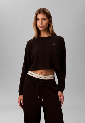 Kvinde iført sort crop top med lange ærmer og sorte bukser med synligt hvidt Calvin Klein-taljebånd mod en ensfarvet grå baggrund.