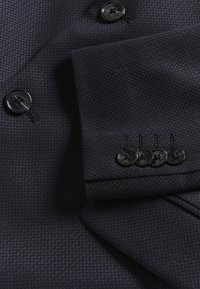 Sötét textúrájú zakó anyag négy fekete ujjgombbal és egy fekete elülső gombbal, közelről látható.