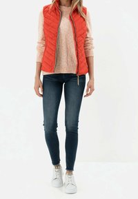 Czerwona pikowana kamizelka ze stójką, noszona na różowym swetrze w cętki, zestawiona z ciemnoniebieskimi dopasowanymi jeansami i białymi trampkami.