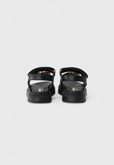 Μαύρα σανδάλια με χοντρή σόλα και διπλή λωρίδα, σε λεία δερμάτινη υφή, διακοσμημένα με αξεσουάρ σε χρυσούς τόνους.