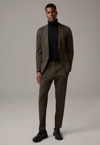 Olivgrüner Anzug mit taillierter Passform, ergänzt durch einen schwarzen Rollkragenpullover. Verfügt über maßgeschneiderte Hosen und schwarze Lederschuhe. Glatte Textur.