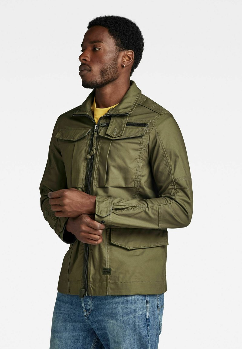 Giacca verde oliva realizzata in materiale leggero, dotata di zip frontale, numerose tasche, colletto alto e dettagli di cucitura a contrasto.