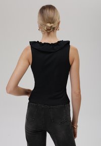 Schwarzes ärmelloses Top mit einem Rüschensaum, aus geripptem Stoff gefertigt. Das Model kombiniert es mit dunklen, engen Jeans. Neutraler Hintergrund.