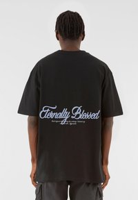 Tricou din bumbac negru cu mâneci scurte și croială largă. Designul de pe spate include textul "Eternally Blessed" în cursiv, de culoare albastru deschis.