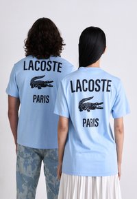 Zwei Personen in hellblauen Lacoste Paris T-Shirts mit schwarzen Krokodil-Logos, die von einem schlichten Hintergrund abgewandt stehen.
