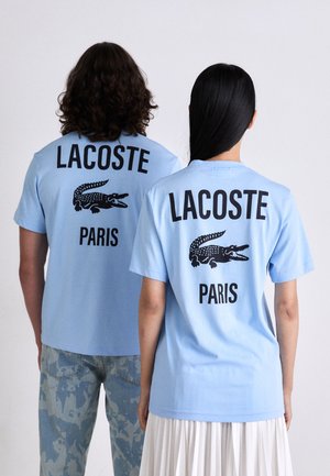 Du žmonės, vilkintys šviesiai mėlynas Lacoste Paris marškinėlius su juodais krokodilo logotipais, atsisukę nugaromis prieš paprastą foną.