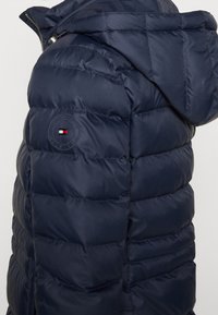 Giacca trapuntata blu navy con cappuccio, realizzata in materiale sintetico, dotata di un design a cassettoni e un logo circolare con accenti rossi e bianchi sul petto.