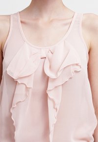 Blouse sans manches rose clair en chiffon, présentant un devant froncé avec un détail nœud au centre, texture douce et tombé fluide.