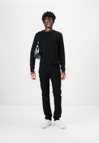 Sort mønstret sweater, sorte bukser og hvide sneakers. Modellen bærer en lille blå og sort taske med en skulderrem. Ensfarvet baggrund.