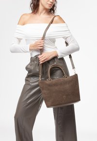 Donna con top bianco a spalle scoperte e pantaloni di pelle grigi, che tiene a tracolla una borsa in suede marrone con manici corti e una tracolla.