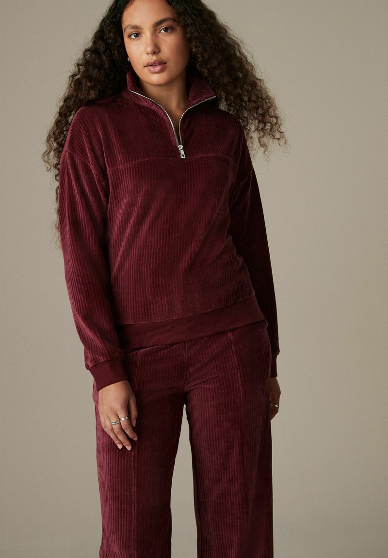 Top con zip bordeaux in corduroy con maniche lunghe, texture a coste e polsini elasticizzati, abbinato a pantaloni coordinati con un design simile.