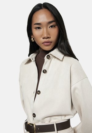 Junge Frau mit langen schwarzen Haaren, die goldene Creolen, eine cremefarbene Jacke mit Knöpfen, einen braunen Rollkragenpullover und einen dunklen Gürtel trägt und in die Kamera schaut.