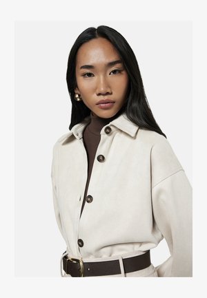 Junge Frau mit langen schwarzen Haaren, die goldene Creolen, eine cremefarbene Jacke mit Knöpfen, einen braunen Rollkragenpullover und einen dunklen Gürtel trägt und in die Kamera schaut.