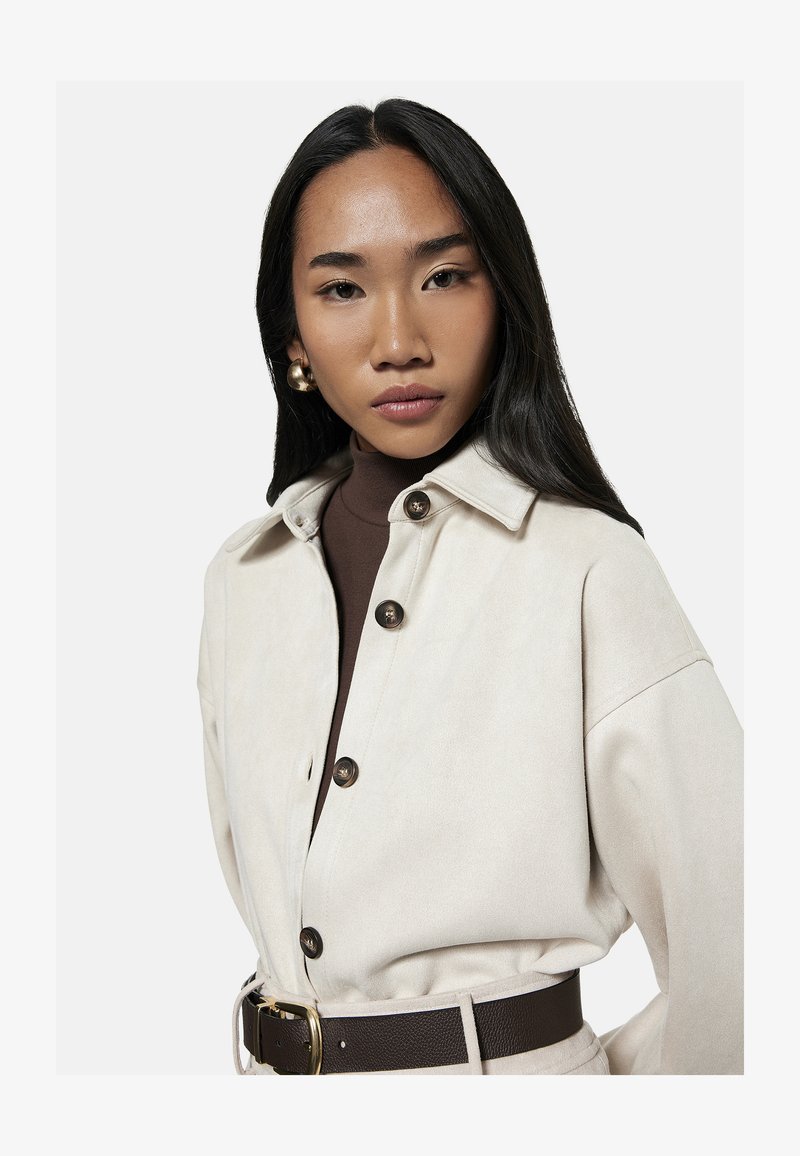 Ung kvinde med langt sort hår iført guldbøjleøreringe, en cremefarvet jakke med knapper, brun rullekrave og mørkt bælte, der kigger på kameraet.