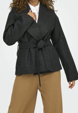 Femme portant un manteau gris foncé ceinturé par-dessus une chemise blanche et un pantalon ample beige côtelé, debout devant un fond clair uni.