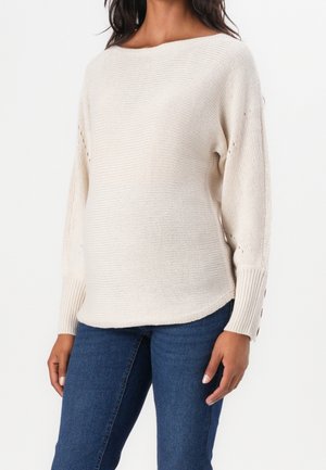 Vrouw draagt een crèmekleurige gebreide trui met knoopdetails op de manchetten en een blauwe spijkerbroek, staand tegen een witte achtergrond.