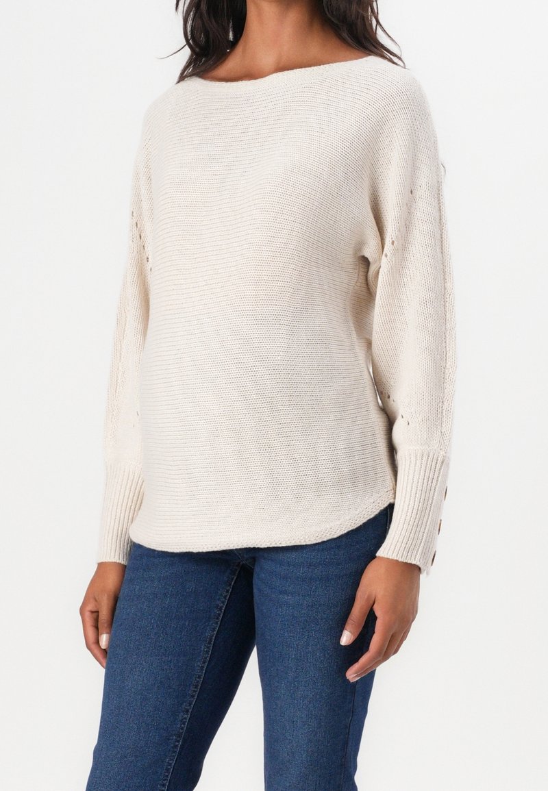 Femme portant un pull en tricot crème avec des boutons aux poignets et un jean bleu, debout devant un fond blanc.