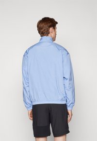 Lys blå, let jakke med høj krave, elastiske manchetter og elastisk talje; glat tekstur; båret over sorte shorts.