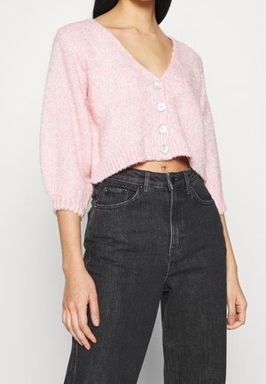 Femme portant un cardigan court rose pâle boutonné avec des manches bouffantes et un jean noir taille haute, debout devant un fond uni.