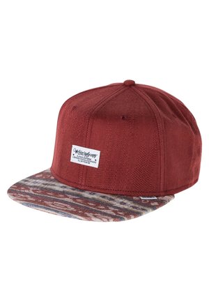 Gorra snapback roja con tejido texturizado y visera plana estampada en tonos tierra, con un parche rectangular blanco con logo en el panel frontal.