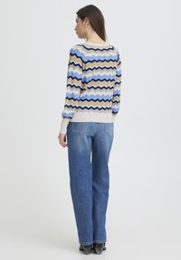 Pull tricoté multicolore avec un motif zigzag en bleu, beige et marron foncé, associé à un jean bleu taille haute.