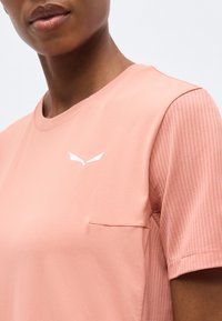 Εγγύς λήψη ενός ατόμου που φοράει πουκάμισο ροδάκινου με κοντά μανίκια με υφή και μικρό λευκό λογότυπο στο στήθος.