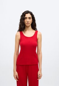 Γυναίκα με μακριά σγουρά μαλλιά που φοράει κόκκινο ριμπ αμάνικο τοπ και αντίστοιχο κόκκινο ριμπ παντελόνι, στέκεται μπροστά σε απλό λευκό φόντο.