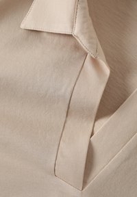 Tela beige clara con una textura suave, que presenta un collar en punta y detalles de costura visibles. El diseño enfatiza líneas limpias y minimalismo.