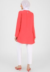 Tunika lunga corallo con maniche lunghe, dotata di un'apertura a chiave sulla schiena, abbinata a pantaloni bianchi a sigaretta e sandali flat rosa.