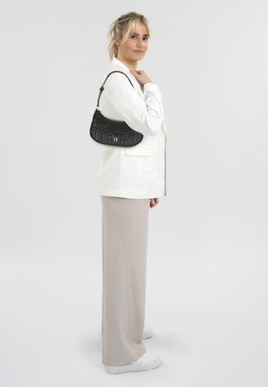 Sac à bandoulière noir à motif avec une finition lisse, porté par une personne en blazer blanc et pantalon beige, debout devant un fond clair.