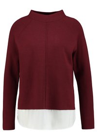 Bordowy sweter z wysokim dekoltem i rękawami raglanowymi, nałożony na białą dolną część. Miękka tekstura i czysty design.