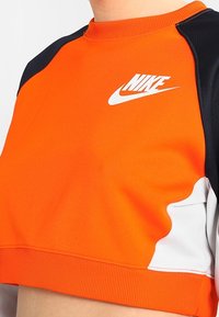 Felpa accorciata Nike realizzata in tessuto arancione brillante, con accenti neri sulle spalle e dettaglio del logo bianco sul petto. Texture liscia.