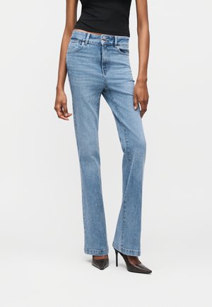 Naine, kes kannab helesiniseid kõrge vöökohaga sirge säärega teksapükse, musta varrukateta topi ja musti teravate ninaga kontsakingi, seisab valge tausta ees.