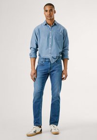 Camicia in denim azzurro chiaro con patta dei bottoni e taschino sul petto; abbinata a jeans slim-fit blu scuro e sneakers bianche con accenti neri.