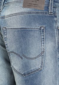 Los jeans de mezclilla de color azul claro presentan un bolsillo trasero con detalles de costura. Una etiqueta de tela con texto es visible cerca de la cinturilla.