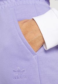 Lila bomullsjoggers med sidoficka och broderad adidas-logga. Vitrandig manschett synlig, vilket visar kontrastdetalj.