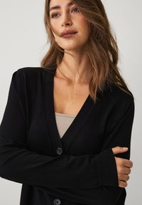 Zwarte gebreide cardigan met een V-hals, lange mouwen en twee knopen aan de voorkant. Het model draagt er een neutrale tanktop onder. Glad textuur.