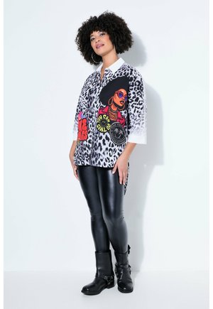 Kvinde med krøllet hår iført leopardprintet skjorte med farverigt motiv, sorte læderleggings og sorte ankelstøvler, stående op ad hvid væg.