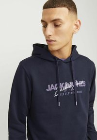 Sudadera azul marino fabricada en un tejido suave con capucha ajustable. Presenta un logotipo en texto blanco en la parte delantera, incluyendo el nombre de la marca y el año de establecimiento.