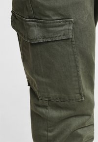 Grüne Cargo-Hosen bestehen aus einem weichen Stoff, haben eine seitliche Tasche mit Klappe und einen lockeren Schnitt. Das Material hat eine glatte, leicht glänzende Textur.